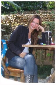 lady sitting at a table with a coffee. she has long brown hair a black jacket and blue jeans. smiling.