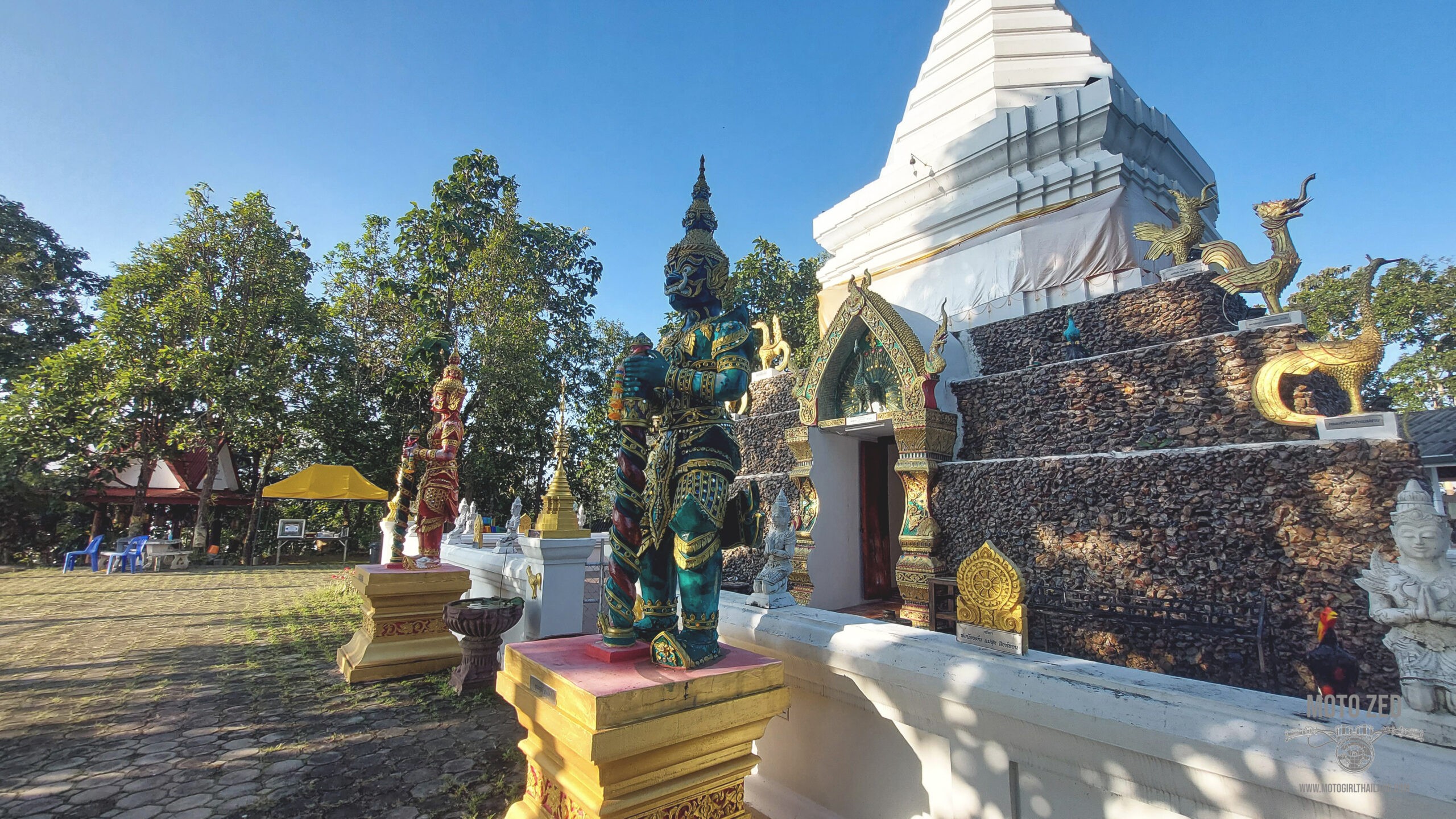 Thai temple with Yak statues in front with a white sky and green trees.