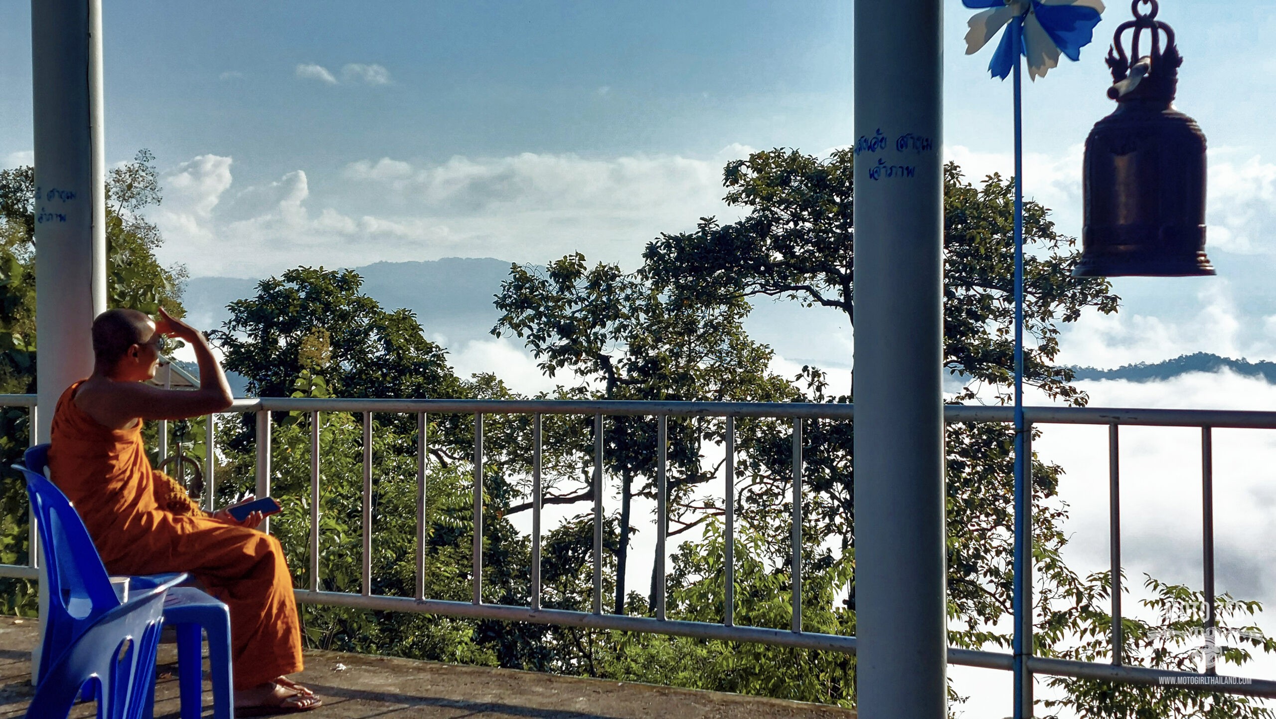 a monk in orange robes looking out to a beautiful sky with clouds and a large temple bell. Samoeng. Chiang Mai, Thailand. Motogirlthailand. Northern Thailand