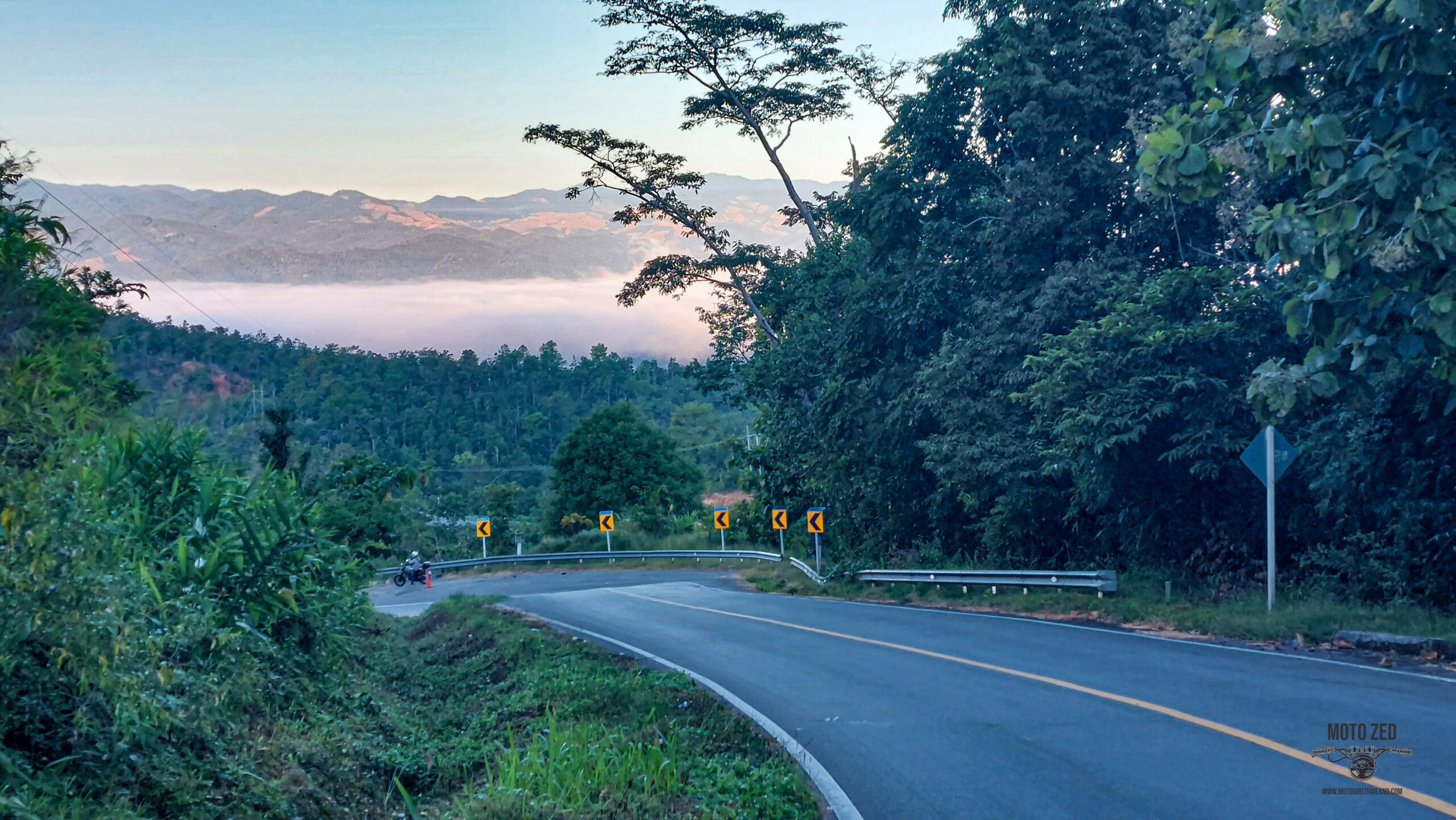 Twisty road surrounded by lush green landscape of trees and a view of mountain with low clouds. Thailand.