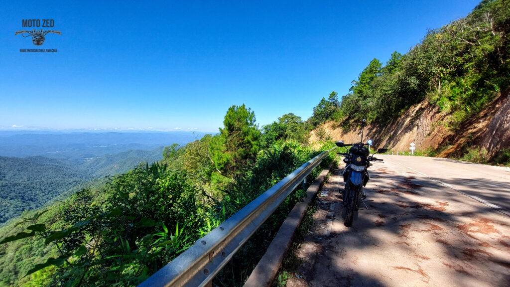 motorcycle parked at a mountain viewpoint with lush green landscape and blue sky. The horizon has many mountains.