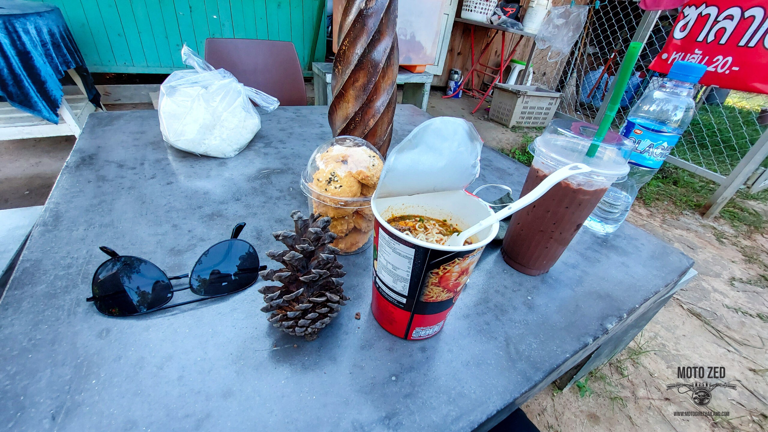 cup noodles, chocolate drink and biscuits sitting on a metal table. There are sunglasses and a bottle of water and a pinecone. In the background are tools and a wire fence